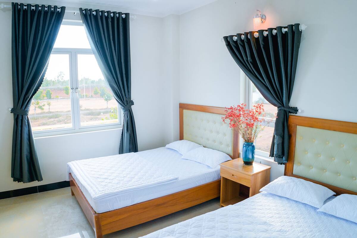 Habitación de hotel con dos camas, una mesita de noche con un jarrón de flores y una ventana con cortinas.