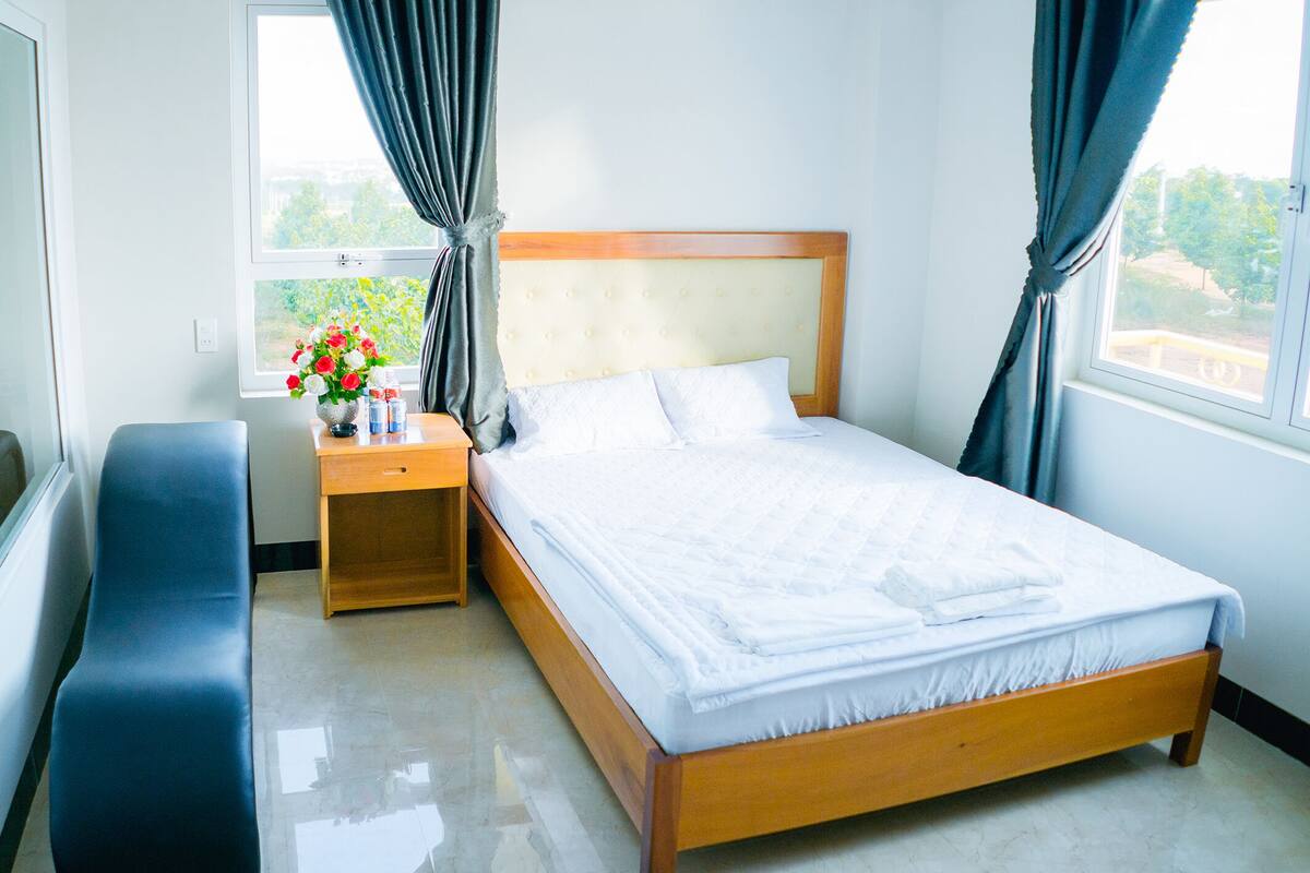 Una cama bien hecha con cabecera de madera, una mesita de noche con un florero y un banco.