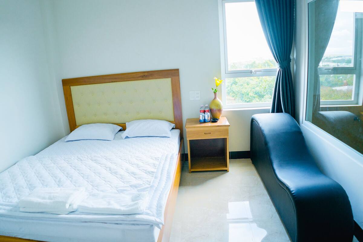 Una cama bien hecha con almohadas, una mesita de noche con un florero y botellas, un banco y una ventana con cortinas.