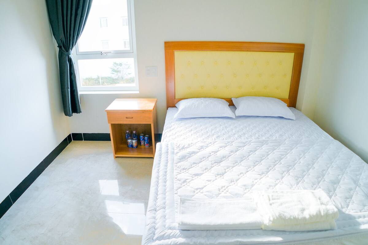 Una cama bien hecha con cabecera de madera, una mesita de noche con botellas de agua y una ventana con cortinas.