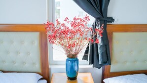 Ein Hotelzimmer mit zwei Betten, einem Holfnachtkästchen, einer Vase mit roten Blumen und einem Fenster mit schwarzen Vorhängen.