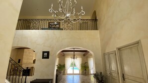 Un hall espacioso con techo alto, una imponente lámpara de araña, una escalera con barandillas ornamentales y vista a una piscina a través de un arco.