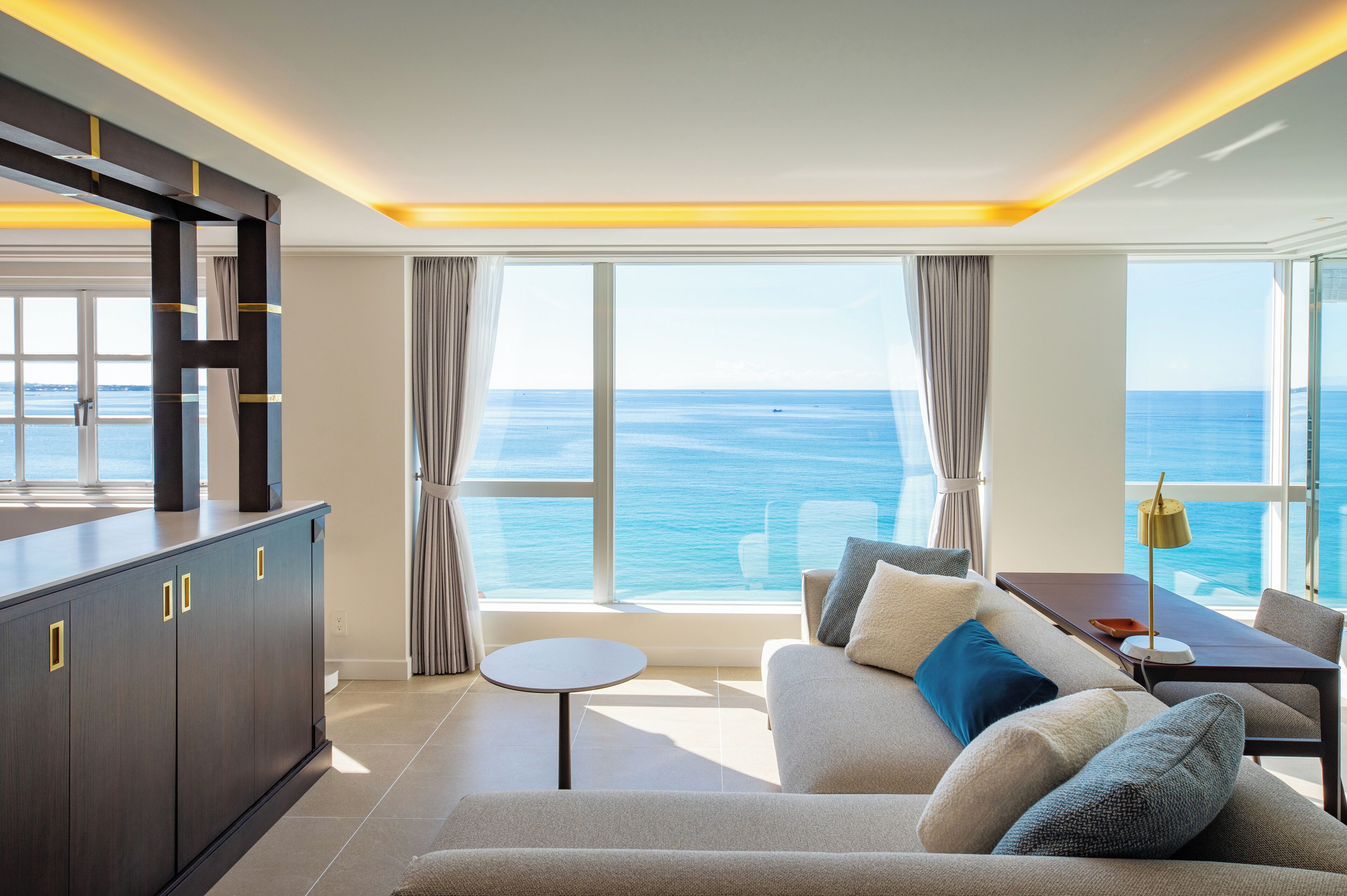 A modern living room with a large window offering a view of the sea, a sofa, a small round table, and a cabinet.
