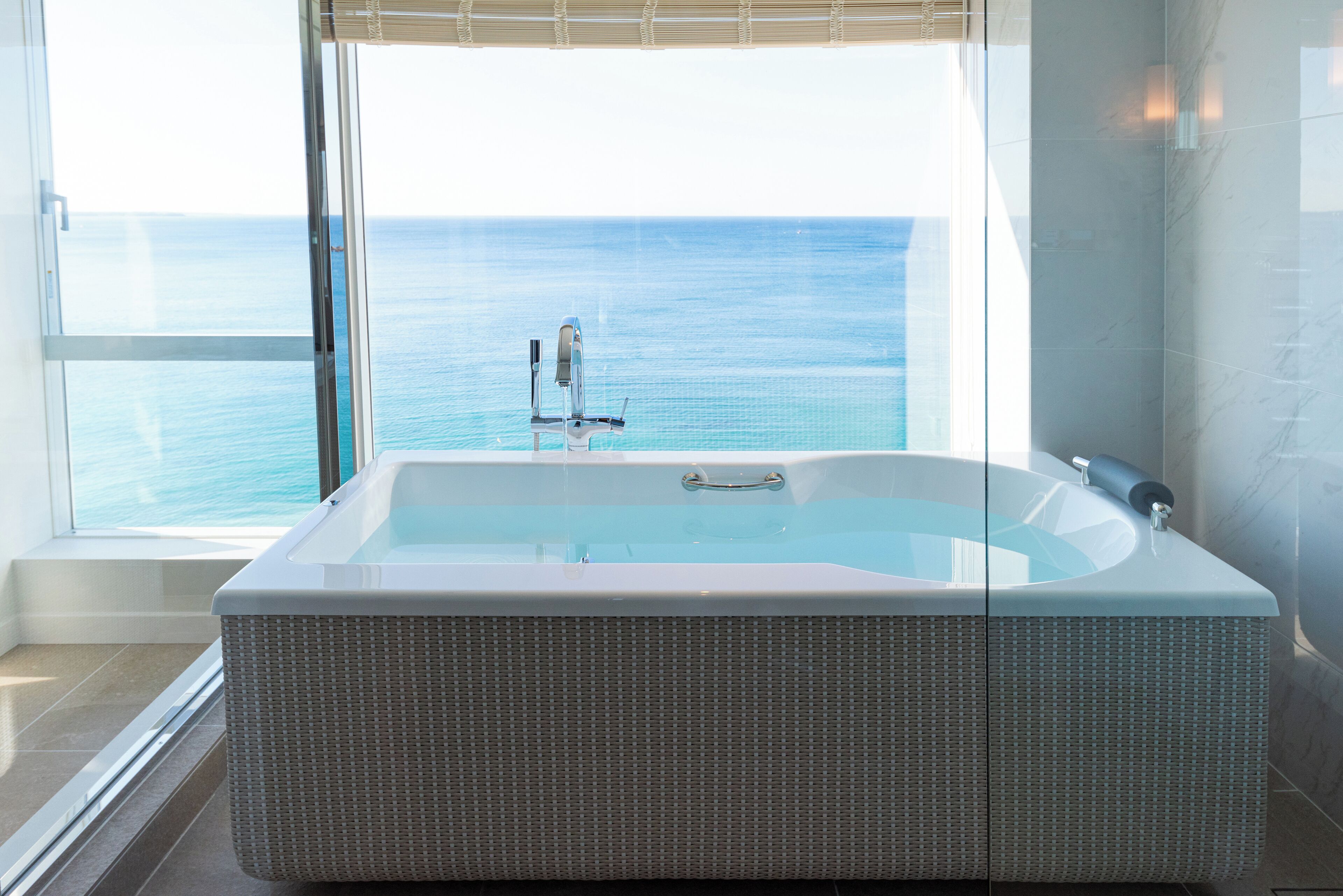 A modern bathroom with a large bathtub, a window with a view of the ocean, and a marble wall.