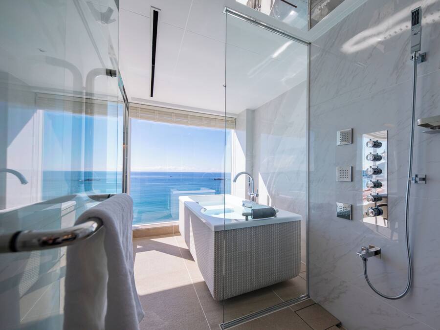 A modern bathroom with a glass-enclosed shower, a bathtub, and a large window offering a sea view.