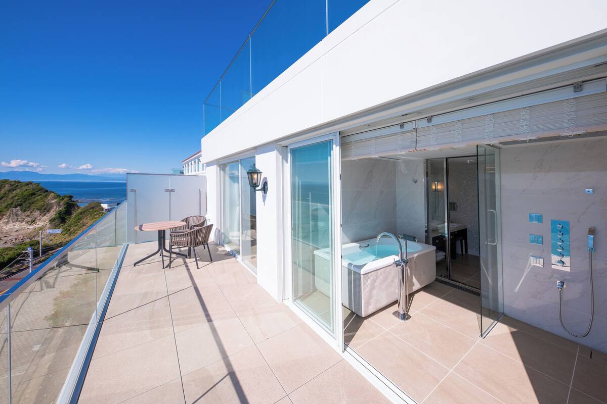 Ein moderner Balkon mit Glasgeländer, einer Sitzecke und einem badezimmerähnlichen Raum mit Badewanne und Dusche.
