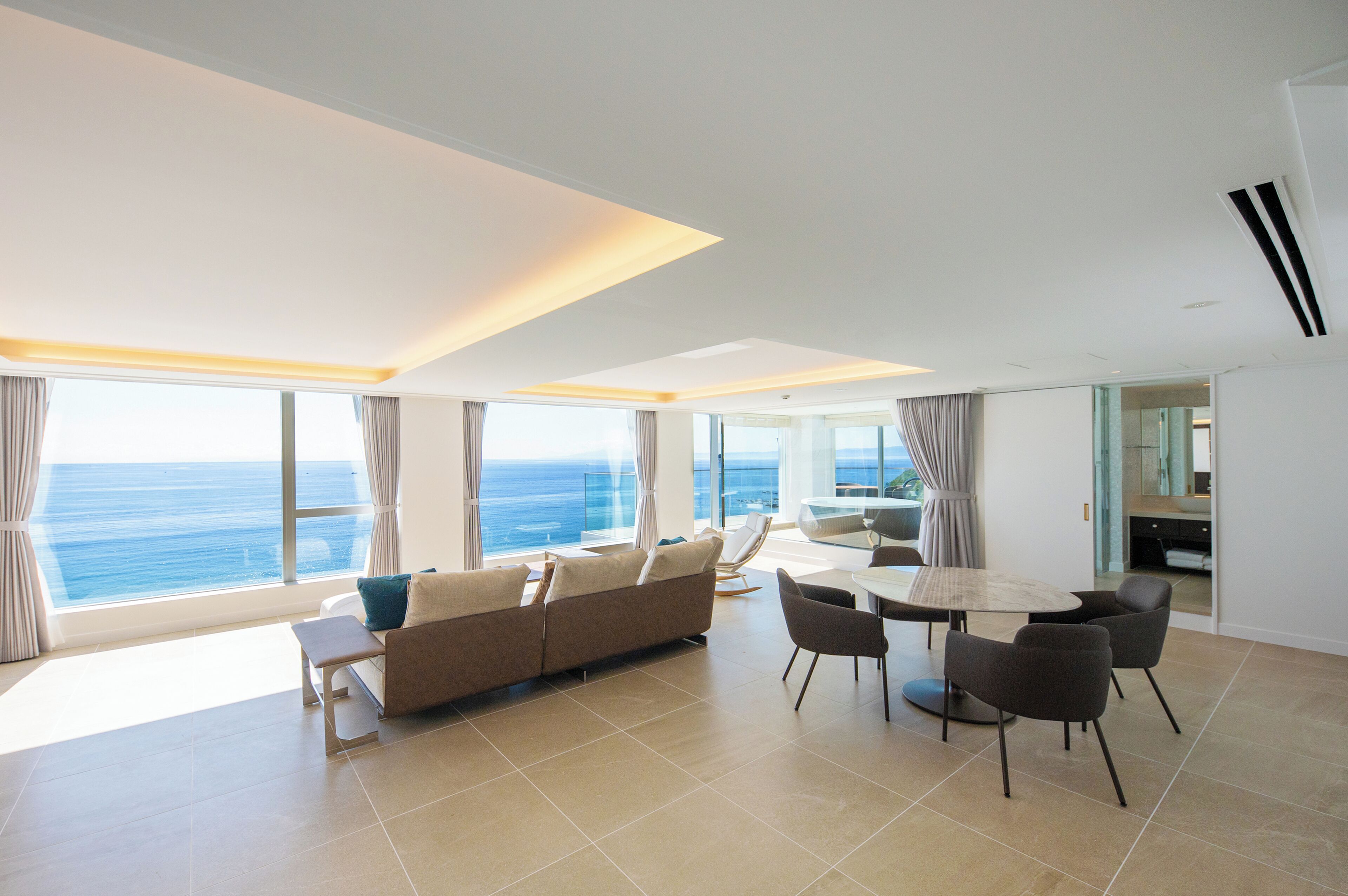 Een moderne woonkamer met een bank, eettafel en stoelen, grote ramen met uitzicht op de oceaan en een keukenhoek.
