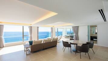 Een moderne woonkamer met een bank, eettafel en stoelen, grote ramen met uitzicht op de oceaan en een keukenhoek.