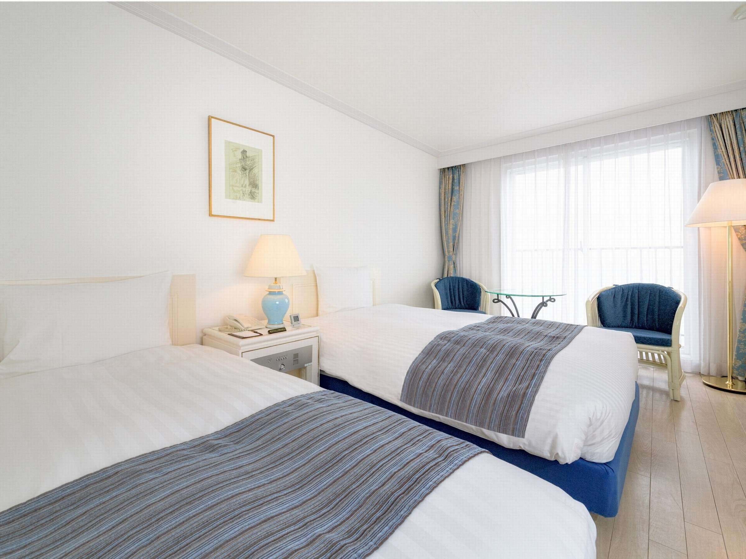 A hotel room with two beds, a bedside table with a lamp, a chair, and a window with curtains.