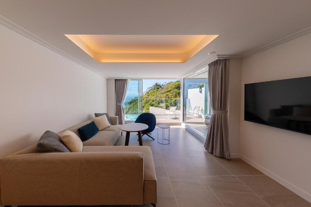 A modern living room with a sofa, a small table, and a TV. There is a balcony with a view of the outdoors.