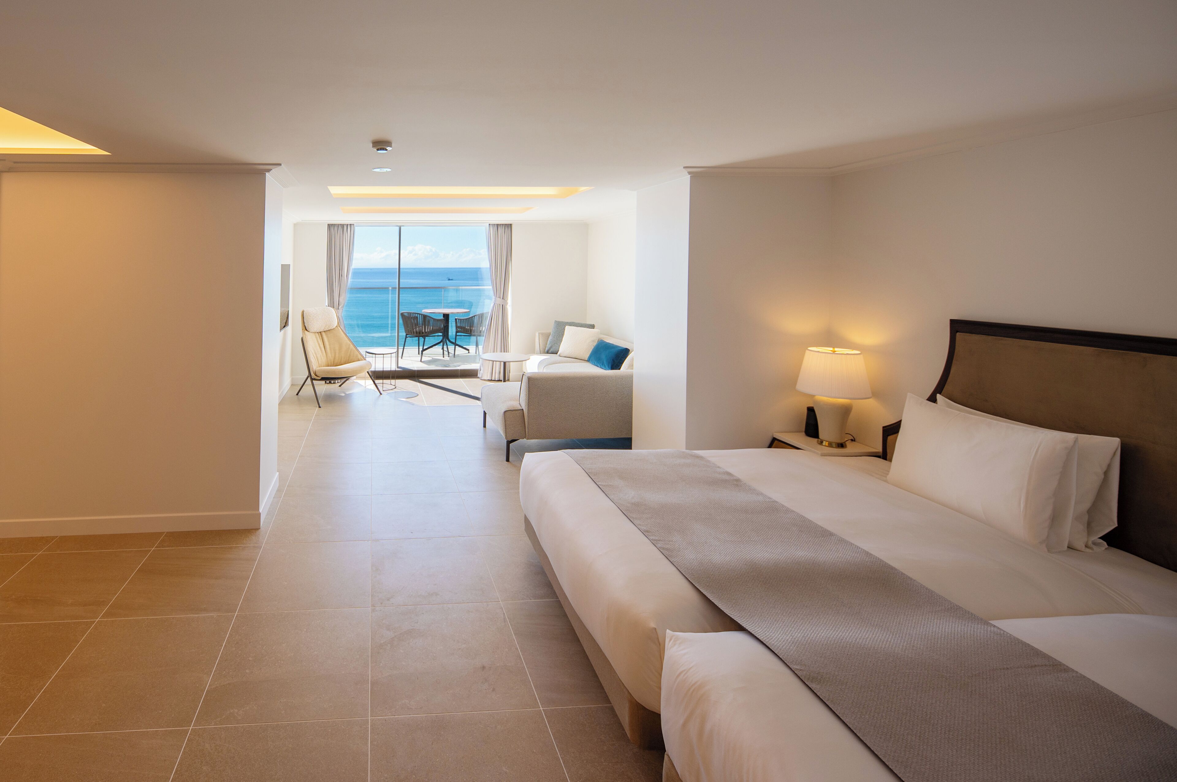 Habitación de hotel con una cama grande, zona de estar con vistas al mar y balcón con mesa y sillas.