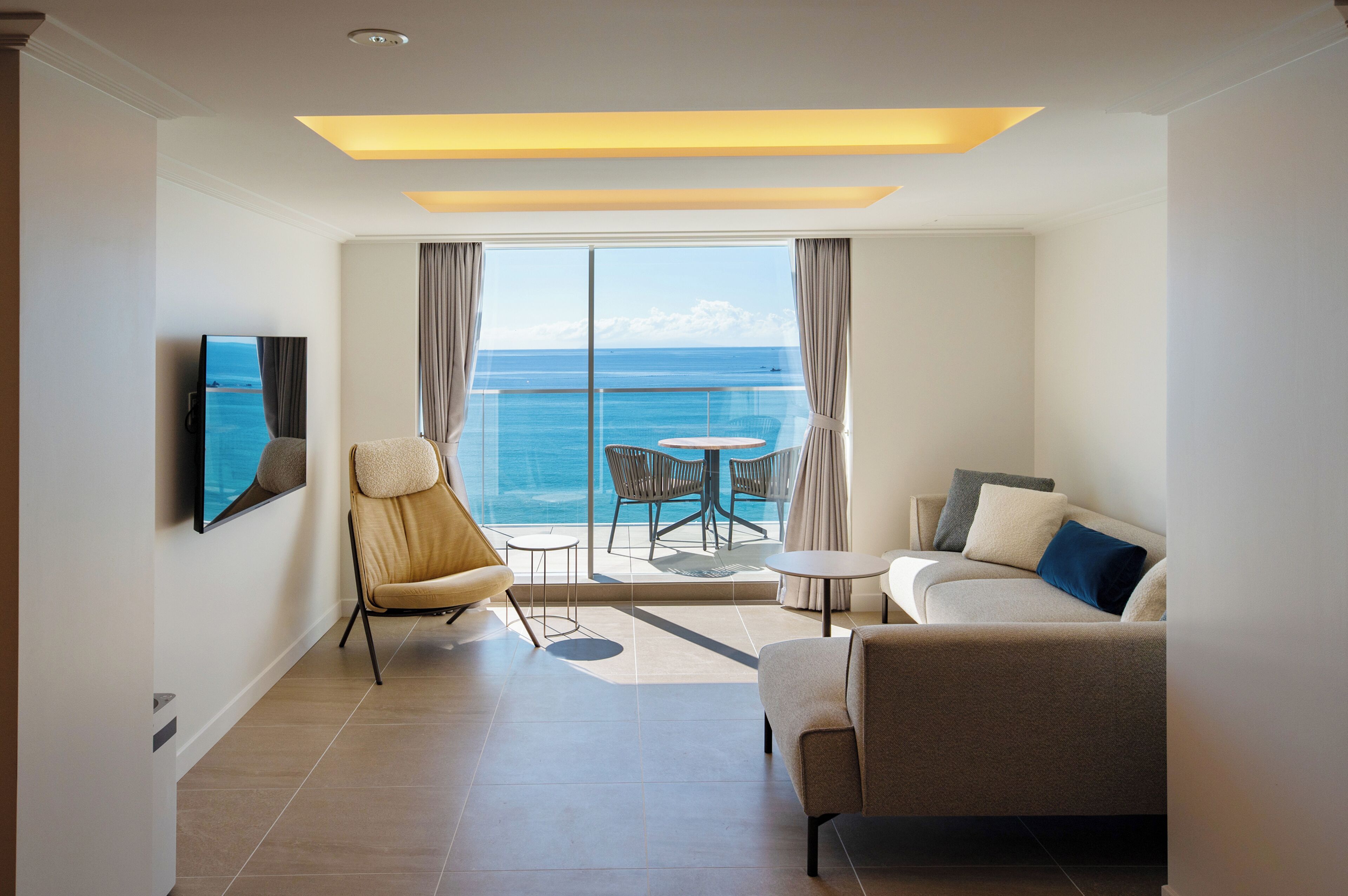 A modern hotel room with a balcony, a sofa, a chair, a small table, and a TV.