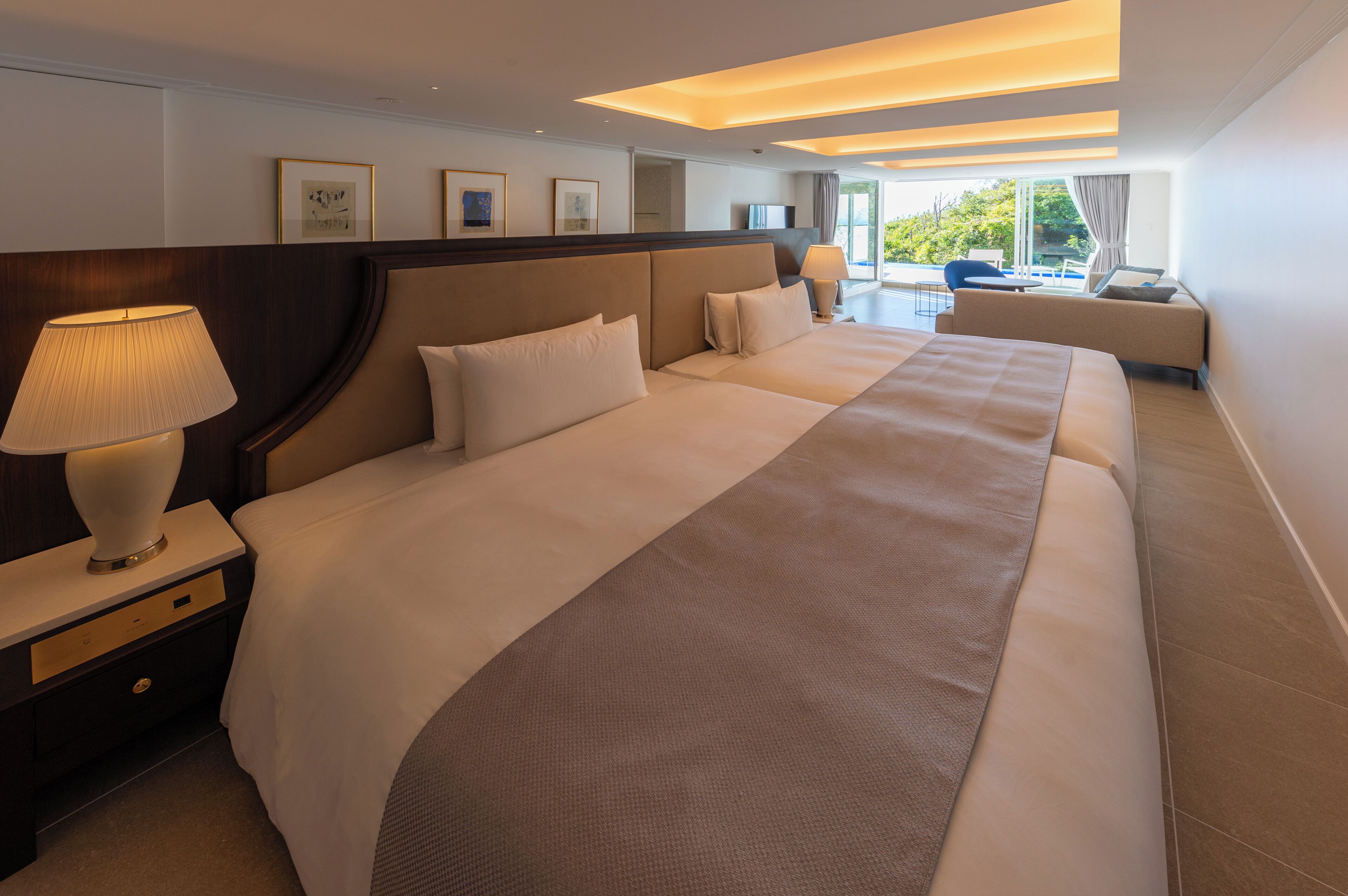 A large, well-appointed hotel room with a long bed, a bedside table with a lamp, and a view of the outdoors.