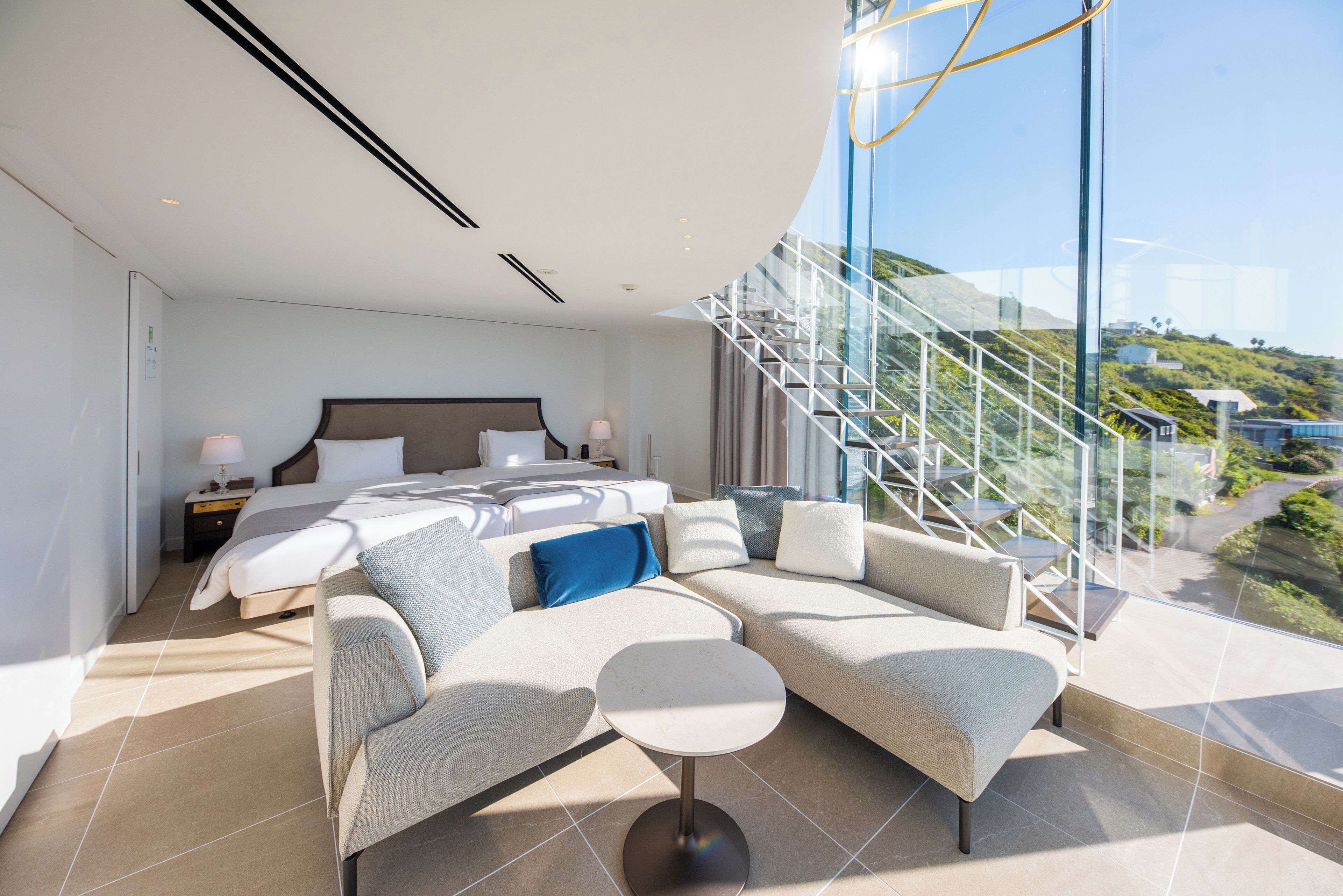Een moderne hotelkamer met een groot bed, een slaapbank, een klein tafeltje en uitzicht op een trap en groen buiten.