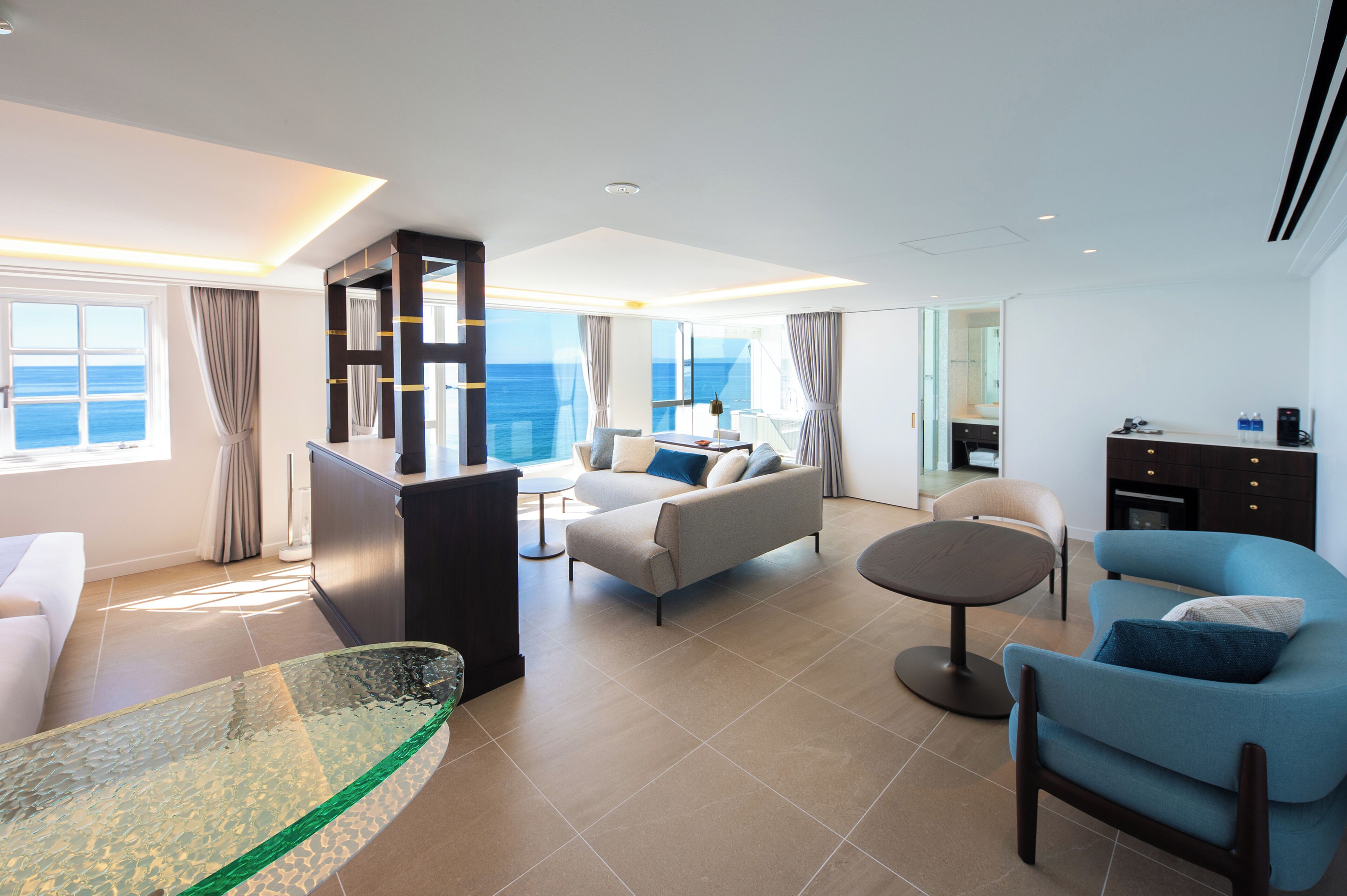 A modern living room with a sofa, armchairs, a glass coffee table, and a view of the ocean.