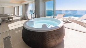 Une suite moderne dotée d’un grand jacuzzi, d’un balcon avec vue sur la mer et d’un coin salon comprenant un canapé et des fauteuils.
