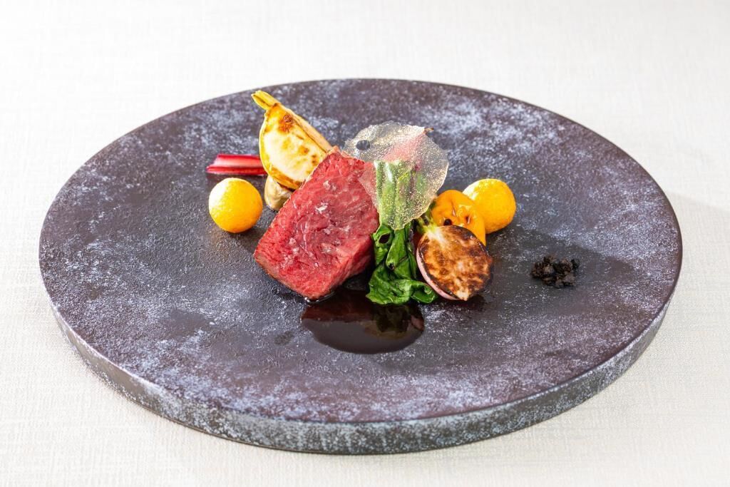 A round, dark-colored plate with a piece of red meat, some vegetables, and a small yellow fruit.