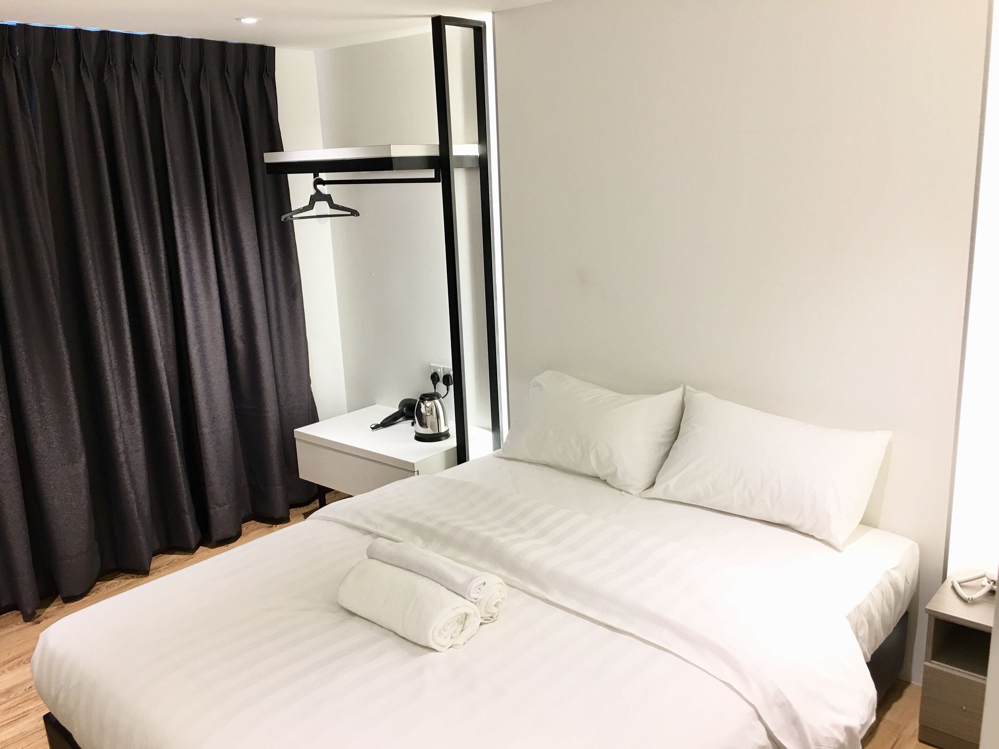 Una camera d'albergo con un letto grande, un comodino con un bollitore, un attaccapanni e una tenda nera.