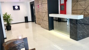 A modern hotel lobby with a reception desk, a digital sign, a trash bin, and a potted plant.