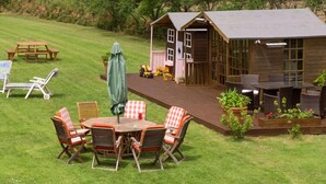Una terraza de madera con mobiliario exterior, un cobertizo pequeño y una mesa de picnic en un área con césped.