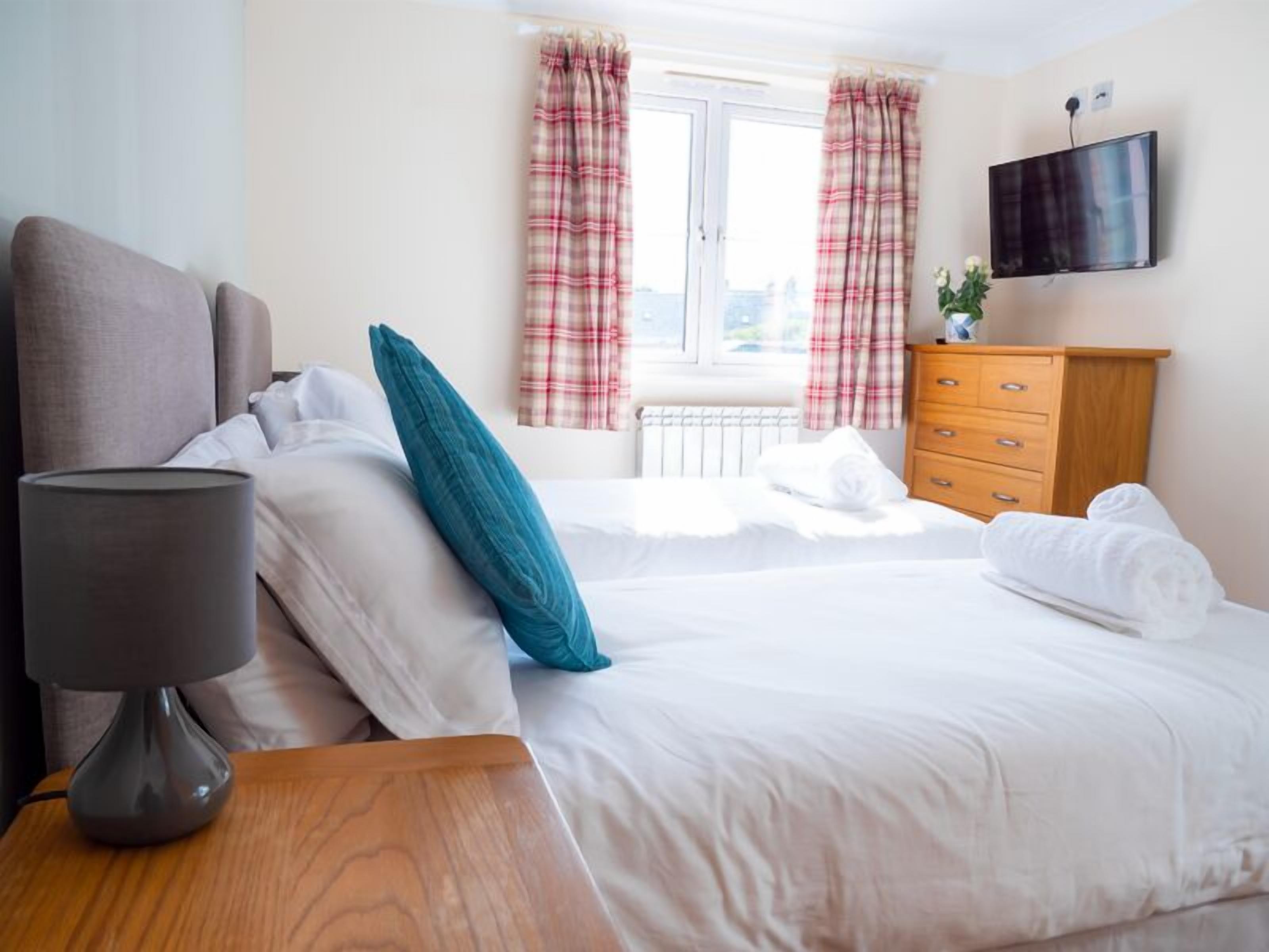 A hotel room with a bed, a bedside table with a lamp, a dresser, a flat-screen TV, and a window with curtains.
