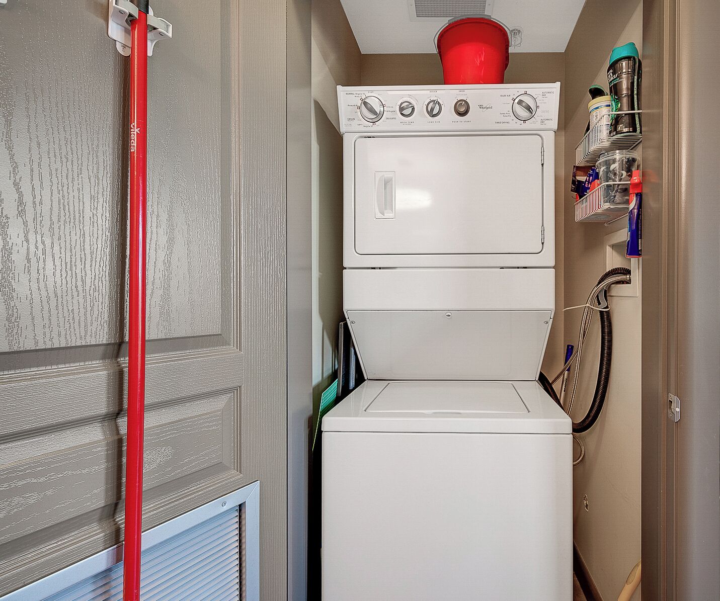 A laundry area with a stacked washer and dryer, a red mop, and a shelf with cleaning supplies.