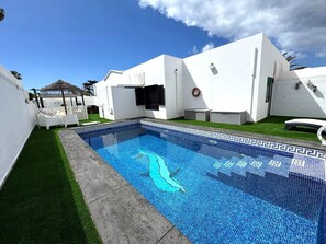 Piscina com padrão geométrico de azulejos, cercada por um edifício branco com porta e janelas, e uma pequena área gramada com cadeiras brancas.