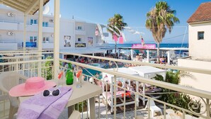 A balcony with a view of a crowded beach area, including a pool and a building with a sign that reads "QUAYSDOLLAGE". - Paralia Studios (Corfu)