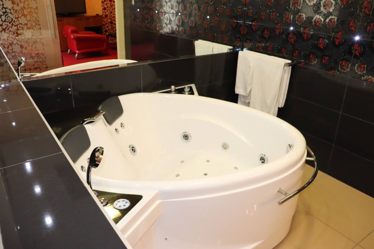 Un baño moderno con una bañera blanca grande, paredes cubiertas de baldosas negras y una silla roja al fondo.