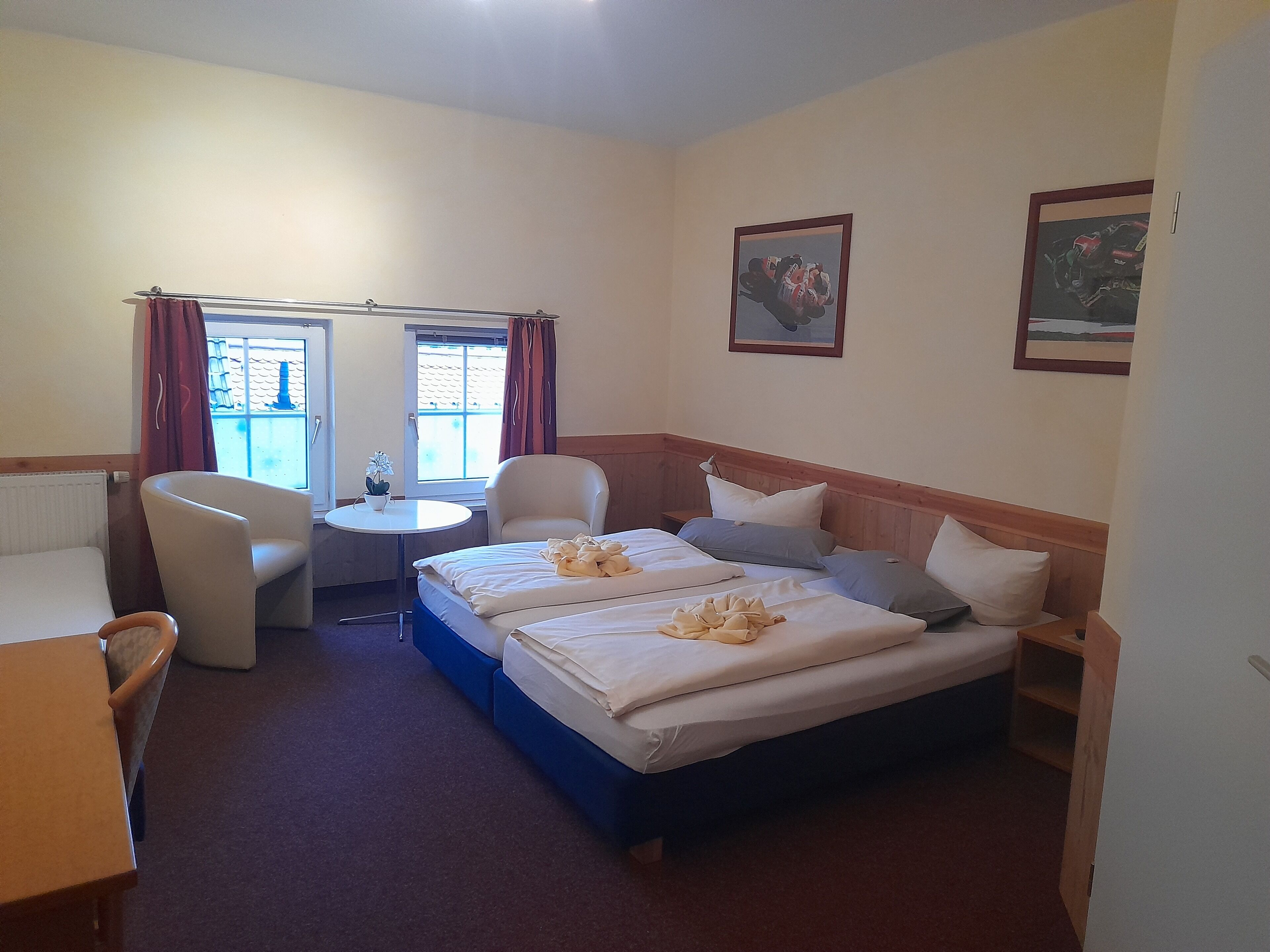 A hotel room with a bed, a desk, a chair, a small table with a plant, and a window with curtains.
