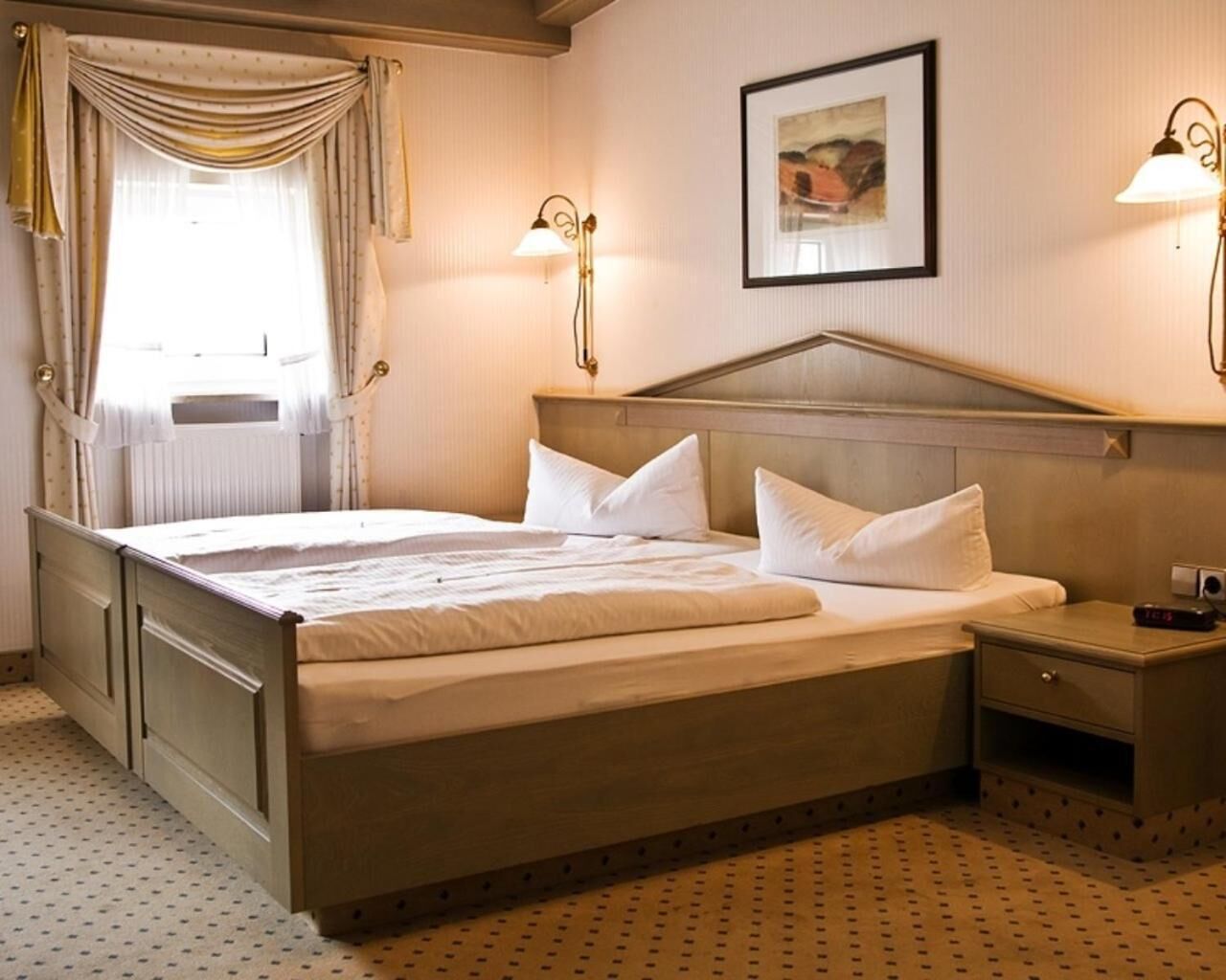 A hotel room with a large bed, two bedside tables, a nightstand, a window with curtains, a wall-mounted lamp, and a framed picture.