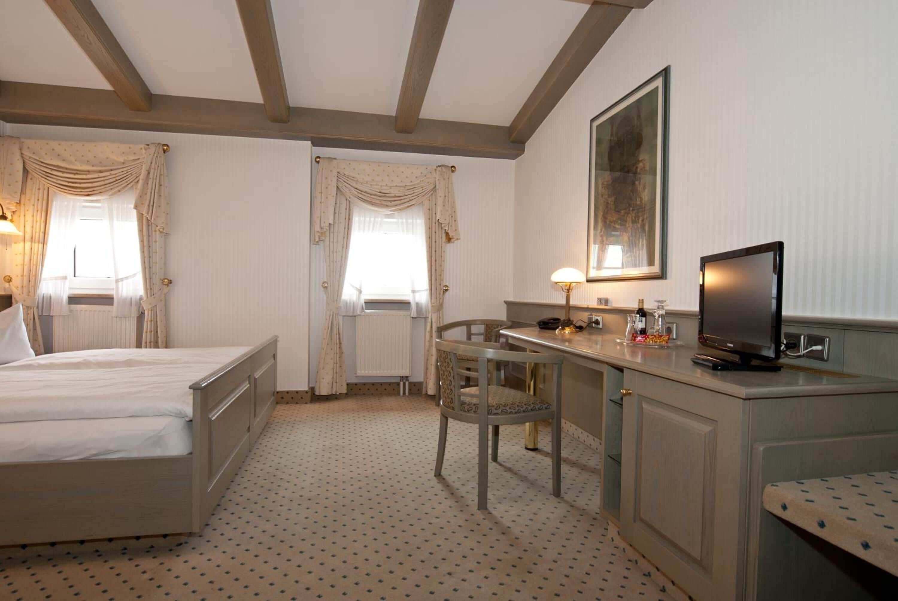 Une chambre d’hôtel comprenant un lit, un bureau avec une chaise, une télévision et une fenêtre avec des rideaux.