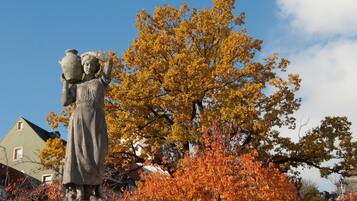 A statue of a woman carrying a pot on her head, set against a backdrop of autumn-colored trees and a clear sky.