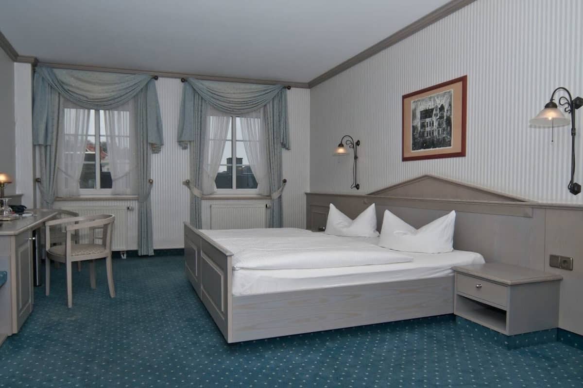 A hotel room with a bed, bedside table, desk, chair, and a window with curtains.