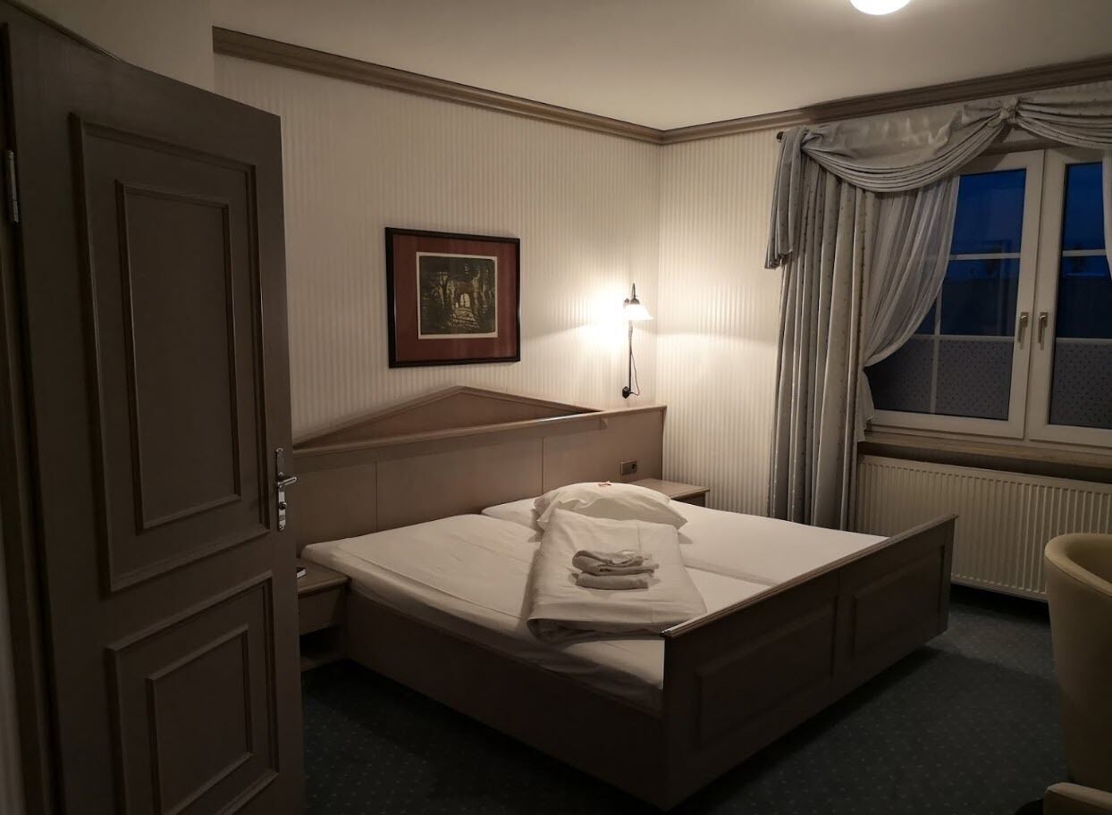Habitación de hotel con cama, mesita de noche, lámpara, un cuadro en la pared y ventana con cortinas.