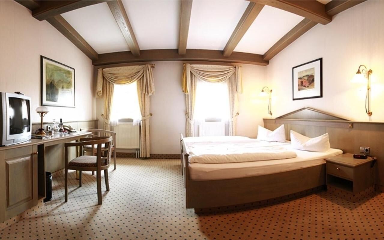 Ein Hotelzimmer mit einem Bett, einem Schreibtisch, einem Sessel, einem Fernseher und zwei Fenstern mit Vorhängen.
