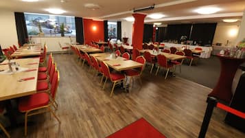Ein Konferenzraum mit mehreren Tischen und Stühlen, großen Fenstern mit Vorhängen und einer Bühne.