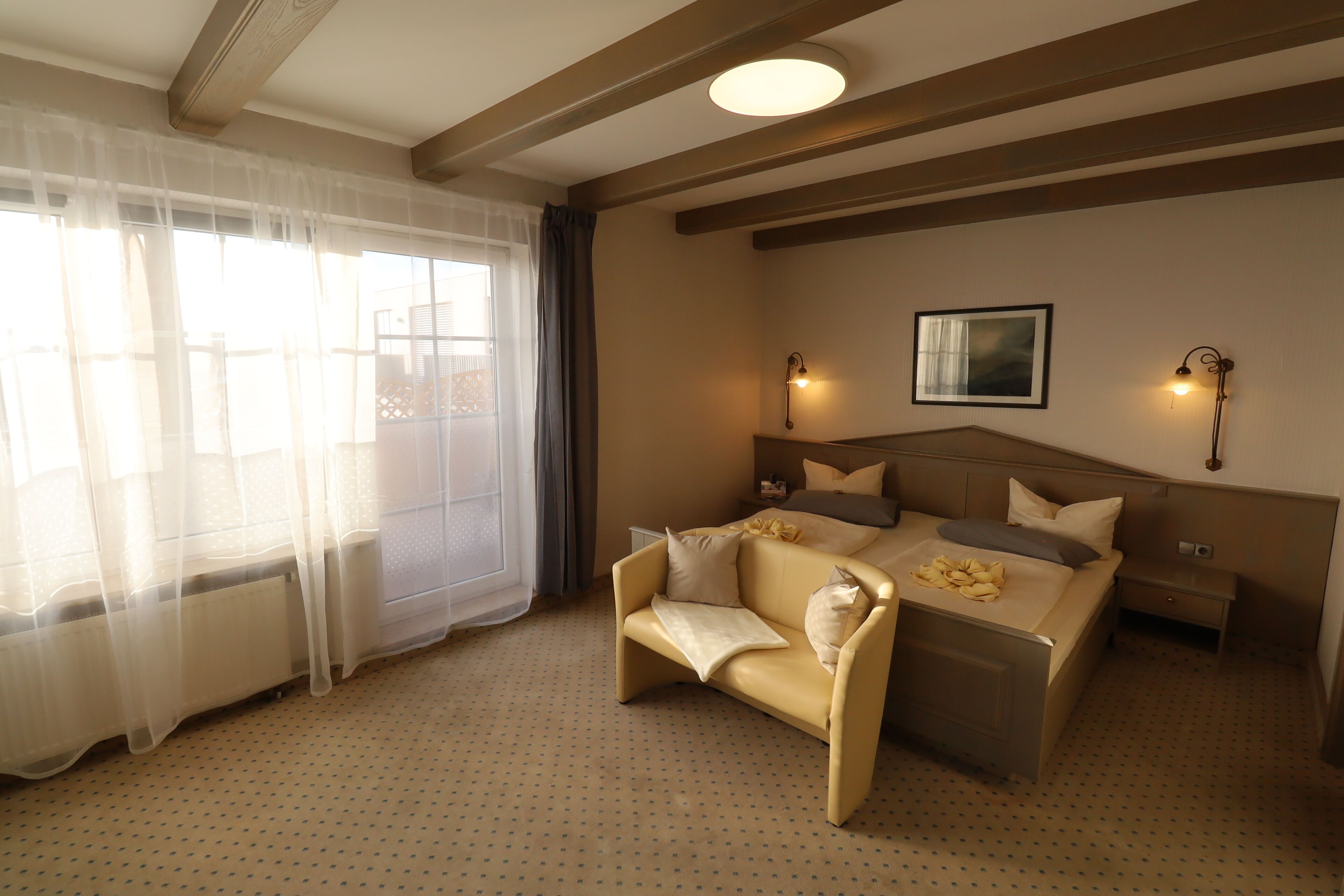 A hotel room with a bed, a sofa, a nightstand, and a window with sheer curtains.