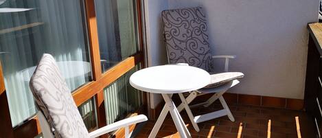 Un balcón con dos sillas blancas y una mesita, una ventana con persianas, y una pared con textura.
