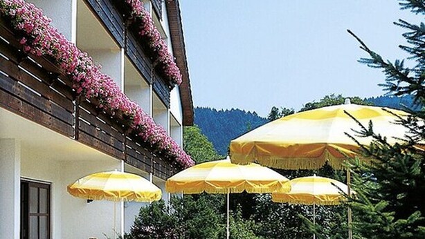 An outdoor patio with white plastic chairs and yellow umbrellas, surrounded by a building with balconies adorned with flowers. - Hotel garni  Zur Weserei (Kandern)