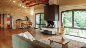 A spacious living room with a stone wall, wooden ceiling beams, a fireplace, and large windows offering a view of greenery. - Telva Alta, country house with tennis in the Dolomites  (Feltre)