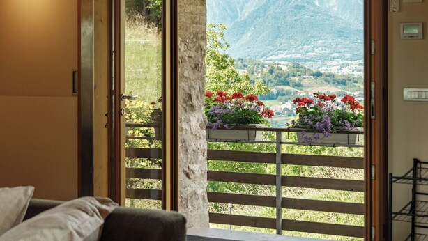 A room with a bed, a wooden wardrobe, and a door leading to a balcony with a mountain view and flower boxes. - Telva Alta, country house with tennis in the Dolomites  (Feltre)