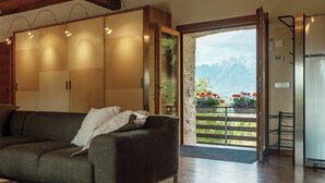A modern living room with a sofa, a wooden ceiling, and a view of mountains through an open door. - Telva Alta, country house with tennis in the Dolomites  (Feltre)