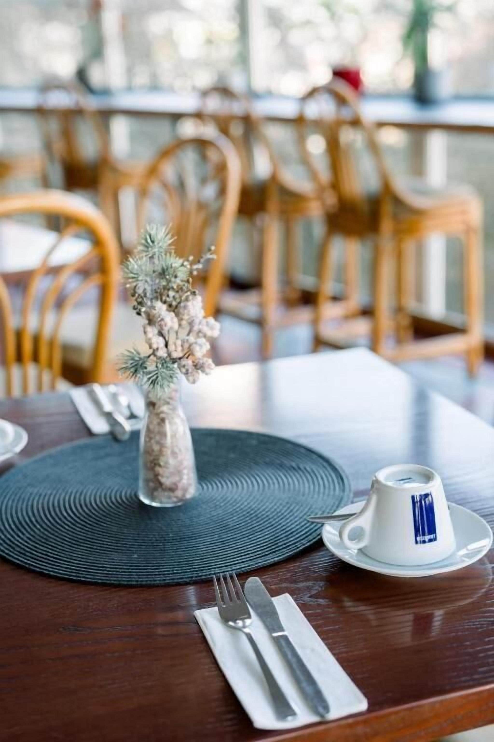 Una mesa de comedor con un camino de mesa, un jarrón pequeño con plantas secas, una taza blanca con un logotipo azul, y un tenedor y cuchillo sobre una servilleta.