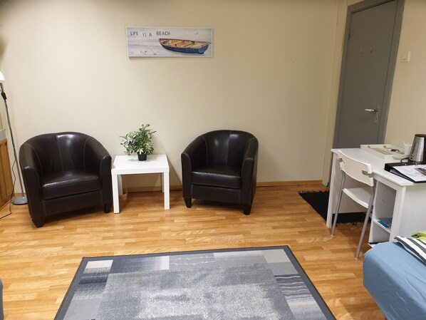 A room with two leather chairs, a small white table with a plant, a desk with a computer, and a bed with a blue bedspread. - Wilhelmsen Romutleie (Vardø)