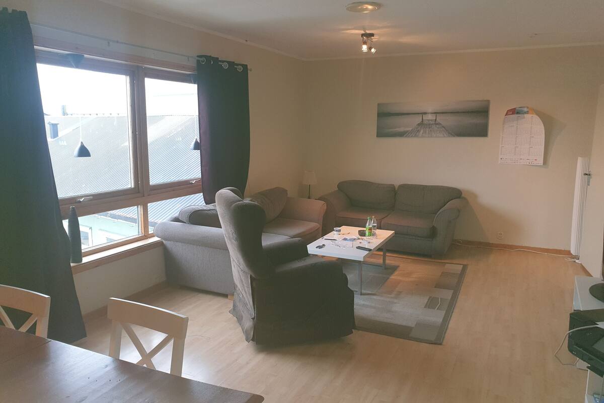 A living room with a sofa, a coffee table, and a dining table. There is a window with curtains, a painting on the wall, and a calendar.