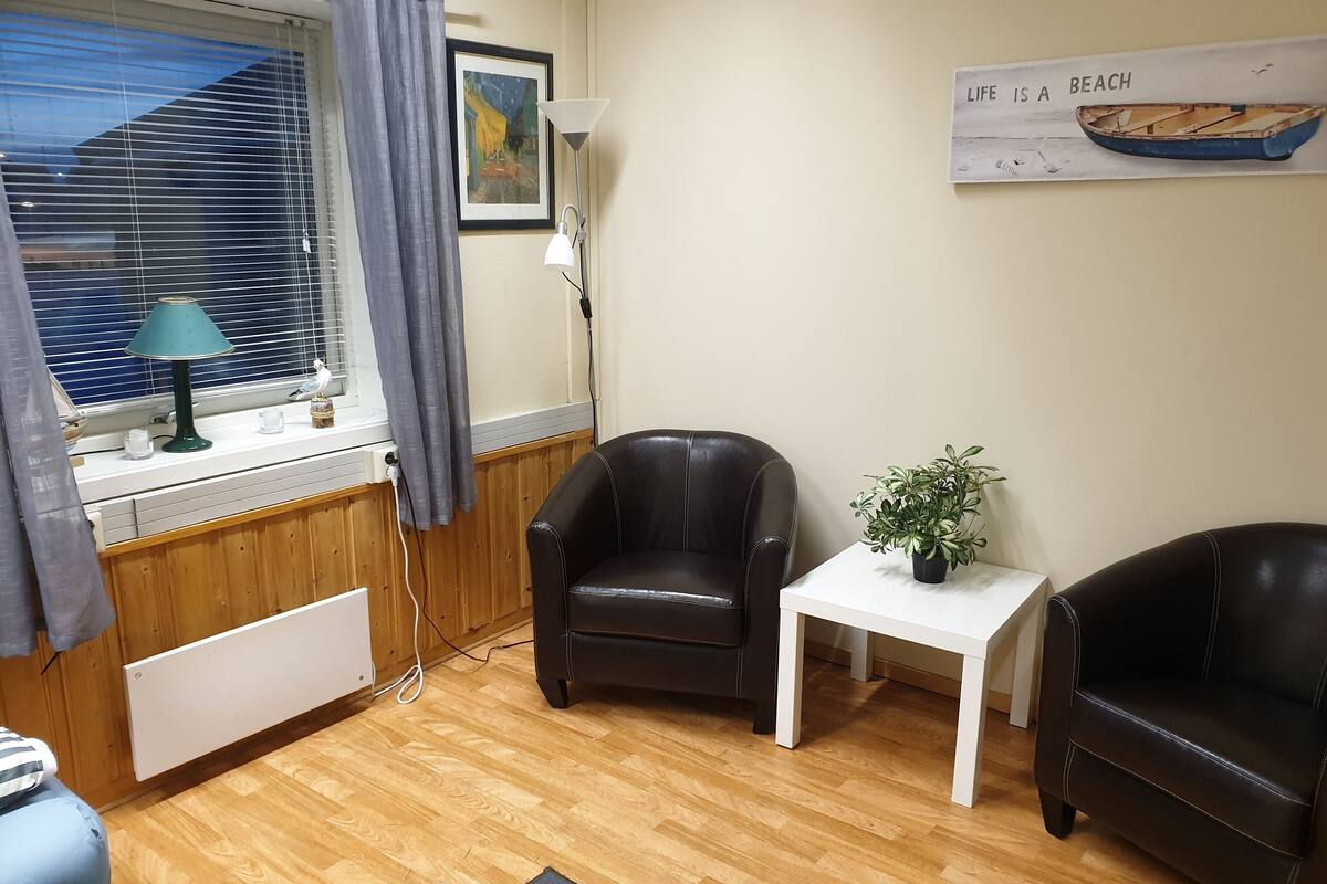 A room with a wooden floor, a black leather chair, a white table with a plant, a framed picture on the wall, and a window with blinds.