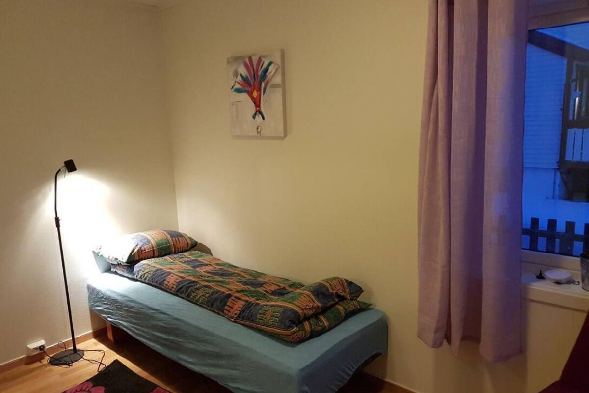 A small, single-bed room with a floor lamp, a colorful bedspread, a wall art piece, and a window with curtains.