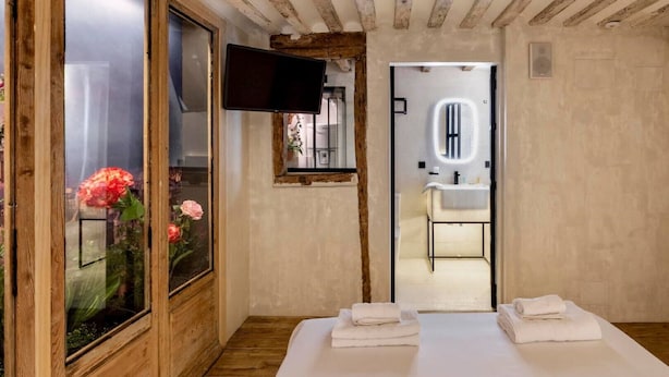Un dormitorio con techo de madera, televisor montado en la pared, ventana con flores y un baño visible a través de una puerta abierta.