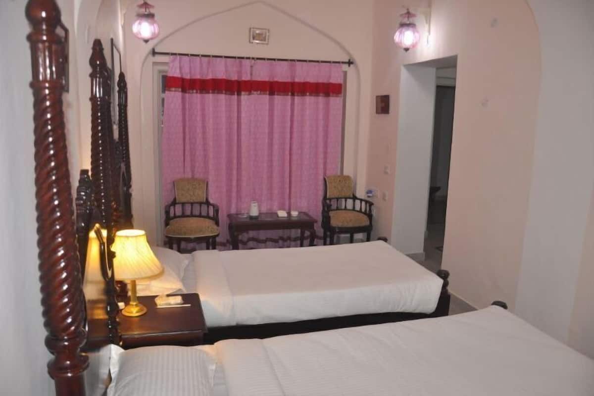 Une chambre d’hôtel avec deux lits, une tête de lit en bois, une petite table avec des chaises, une lampe et une fenêtre avec des rideaux.